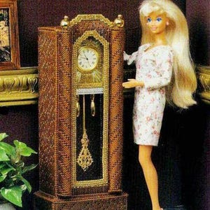 Puede incluir: Un reloj de pie de madera marrón con una esfera dorada y una esfera blanca. El reloj tiene un péndulo y una parte superior decorativa. Una muñeca está de pie junto al reloj.