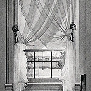 May include: A black and white photo of a window with white curtains. The curtains are tied back with decorative tassels. The window is open and there is a view of a building outside.