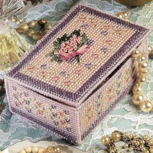 Op de afbeelding: Een rechthoekige, handgemaakte doos met een scharnierend deksel. De doos is voornamelijk lichtroze met een bloemenpatroon en een donkerpaarse rand. Een bloemenboeket met een roze strik siert het deksel. De doos wordt waarschijnlijk gebruikt voor het opbergen van sieraden of kleine souvenirs.