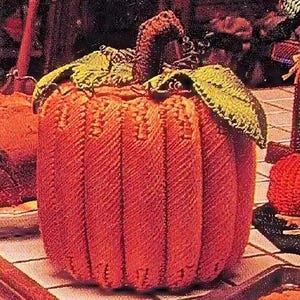 May include: A knitted pumpkin with a textured orange surface and a green stem. The pumpkin is sitting on a white tiled surface.