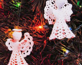 May include: Two handmade, white crochet angel ornaments hanging on a Christmas tree. The angels have detailed wings, halos, and flowing gowns. The tree is decorated with colorful lights, creating a festive holiday scene.