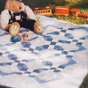 Op de afbeelding: Een wit en blauw gestikt deken met een ruitpatroon. Een teddybeer met een matrozenpet en een overall zit op het deken met een speelgoedtrein en houten blokken.