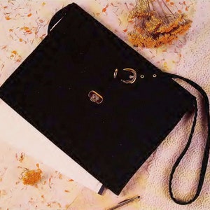 May include: A black velvet pouch with a gold clasp and a black drawstring. The pouch is lying on a white surface with dried flowers.