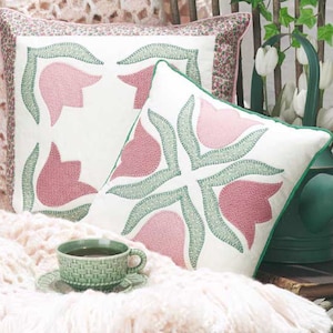 May include: Two white pillow covers featuring pink and green embroidered tulips. The pillows are resting on a white blanket with a green teacup and saucer on top.