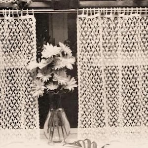Puede incluir: Dos cortinas blancas de ganchillo con un patrón floral cuelgan en una ventana. Las cortinas están hechas de un diseño delicado y calado. Un jarrón de flores blancas se encuentra sobre una mesa frente a la ventana.