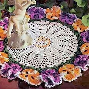 May include: A cream-colored, crocheted doily with a central floral design, surrounded by a border of crocheted pansies in shades of purple, orange, and yellow. A small, off-white cherub statue sits atop the doily.