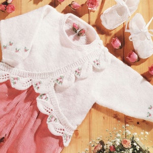 May include: White knitted baby cardigan with a scalloped edge and embroidered flowers. The cardigan is laid out on a wooden surface with pink baby pants and white shoes.