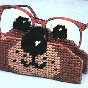 May include: A brown and tan crocheted glasses holder shaped like a dog's face with black eyes and a black nose. The holder is holding a pair of brown glasses.
