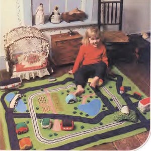 以下が含まれることがあります： 緑と青の道路と線路のデザインのプレイマット。子供がマットの上でミニカーで遊んでいます。