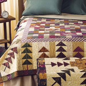 Op de afbeelding: Een patchworkdeken met een bruin, groen en crème kleurenschema met een patroon van gestileerde dennenbomen. De deken ligt op een bed met een groen kussen en een bruin houten hoofdeinde.