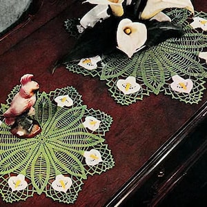 Könnte beinhalten: Zwei grüne Häkeldeckchen mit weißen und gelben Blumendesigns. Die Deckchen liegen auf einer dunklen Holzoberfläche mit einer weißen Keramikvogelfigur in der Mitte eines Deckchens.