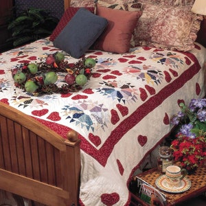 May include: A white and red quilt with a heart pattern is on a wooden bed frame. The quilt has a red border with white hearts. There are pillows on the bed with a floral pattern. A wreath of fruit is on the bed.