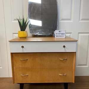 May include: A vintage wooden dressing table with three drawers and black legs. The dressing table has a white top and gold handles. A mirror and a sign that says "My Vintage Market" are on top of the dressing table.