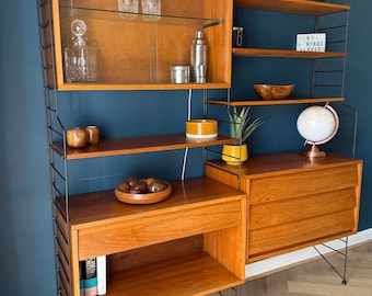 May include: A modular teak wall unit featuring black metal ladder-style supports, a glass-fronted cabinet, open shelving, and two lower drawer units. The shelving displays various decorative items, including glassware, a globe, and a small sign.