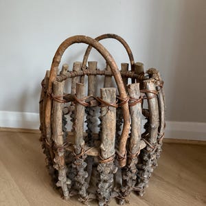 May include: A rustic basket constructed from vertical wooden branches and twigs, featuring two arched handles at the top. The structure is held together by thin, dark brown woven vines that wrap around the textured, weathered wood pieces.