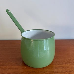 May include: A sage green enamel saucepan with a white interior and a long, slender handle. The pot features a rounded body and a small hole at the end of the handle for hanging. The exterior surface is smooth and glossy with a consistent green finish.