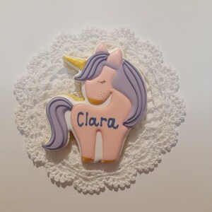 May include: A unicorn-shaped cookie with pink, purple, and gold details, resting on a white, lacy doily. The cookie has the name "Clara" written in blue script. The unicorn has closed eyes and a golden horn.