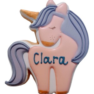 : A unicorn-shaped biscuit with a pink body, purple mane and tail, and a golden horn. The biscuit has the name "Clara" written in blue.