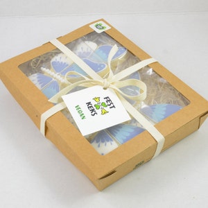 Op de afbeelding: Een doos met veganistische vlinderkoekjes, vastgebonden met een wit lint. De doos is gemaakt van bruin karton en heeft een transparant venster aan de bovenkant. De koekjes zijn versierd met blauwe en witte glazuur. De doos heeft een label met de tekst "FEST KEKS VEGAN".