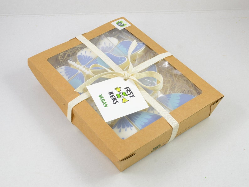 Op de afbeelding: Een doos met veganistische vlinderkoekjes, vastgebonden met een wit lint. De doos is gemaakt van bruin karton en heeft een transparant venster aan de bovenkant. De koekjes zijn versierd met blauwe en witte glazuur. De doos heeft een label met de tekst "FEST KEKS VEGAN".