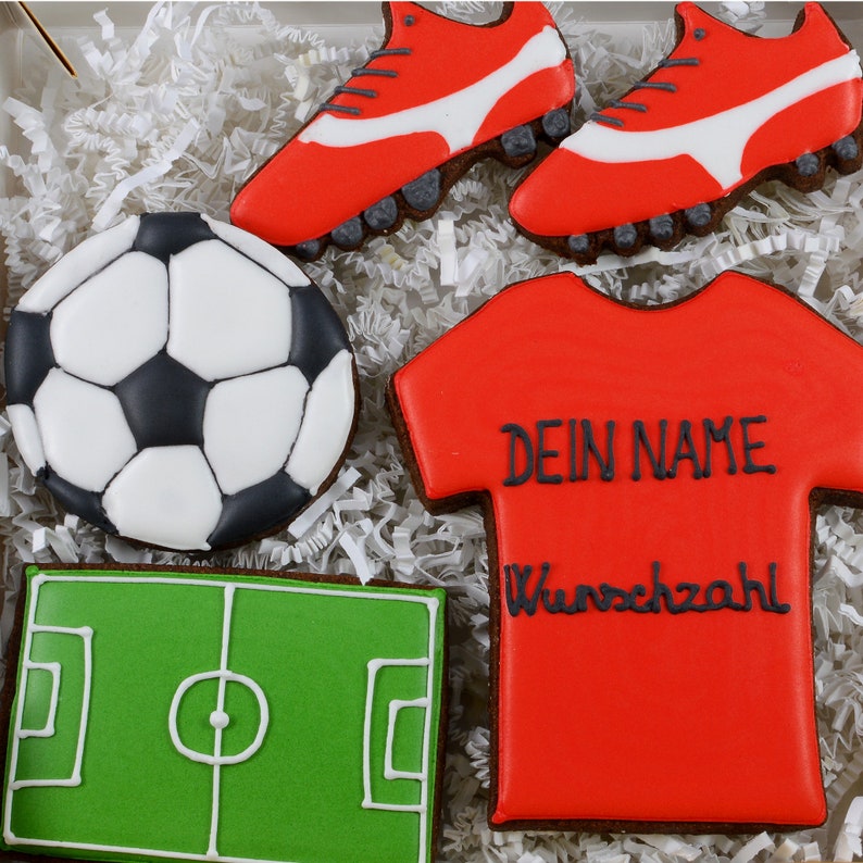 May include: A collection of decorated sugar cookies in a box. The cookies include a soccer ball, a soccer field, a red jersey with text, and two red soccer cleats with white accents.
