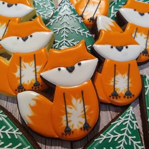 May include: A collection of decorated sugar cookies featuring orange foxes and green trees. The cookies are decorated with white icing and have a snowflake design.
