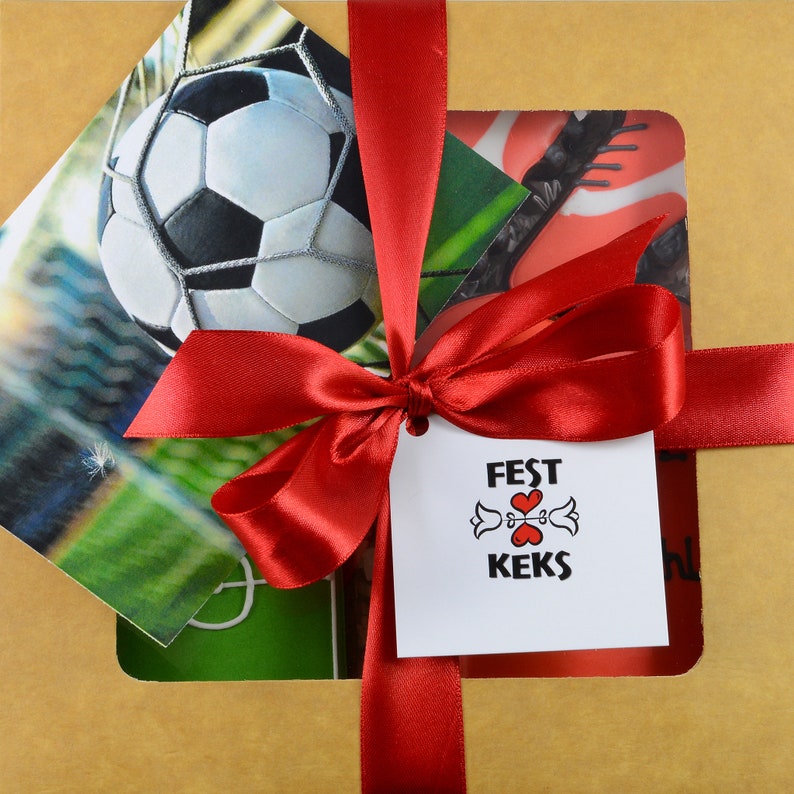 May include: A gift box wrapped in tan paper, tied with a red satin ribbon and bow. The box contains images of a soccer ball and soccer cleats. A white tag with the words "FEST KEKS" is attached to the bow.