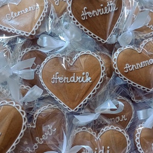 May include: A collection of heart-shaped gingerbread cookies, each individually wrapped in clear cellophane and tied with a white ribbon. The cookies are decorated with white icing and have names written on them, including "Clemens", "Tanja", "Henriette", "Hendrik", "Robert", "Heidi", and "Anna".