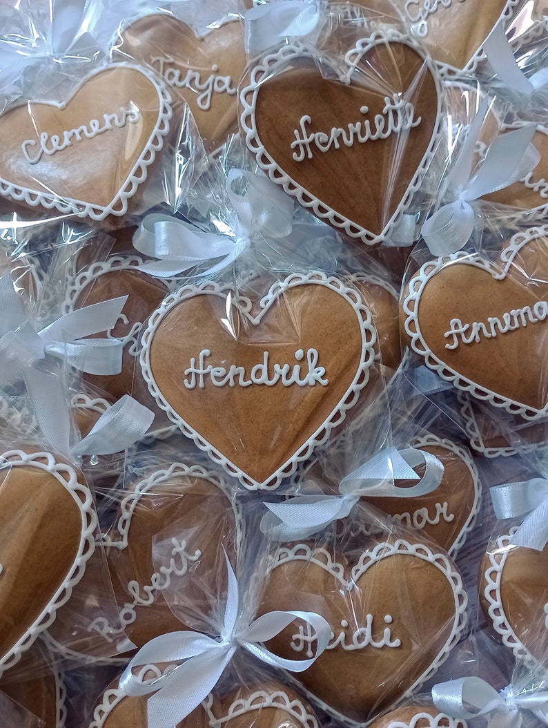 May include: A collection of heart-shaped gingerbread cookies, each individually wrapped in clear cellophane and tied with a white ribbon. The cookies are decorated with white icing and have names written on them, including "Clemens", "Tanja", "Henriette", "Hendrik", "Robert", "Heidi", and "Anna".