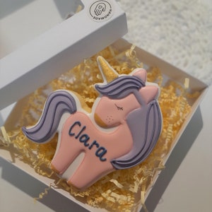 May include: A unicorn-shaped cookie with pink, purple, and gold details, personalized with the name "Clara" in blue. The cookie is presented in a white box with yellow paper filler, and a "HANDMADE" sticker.