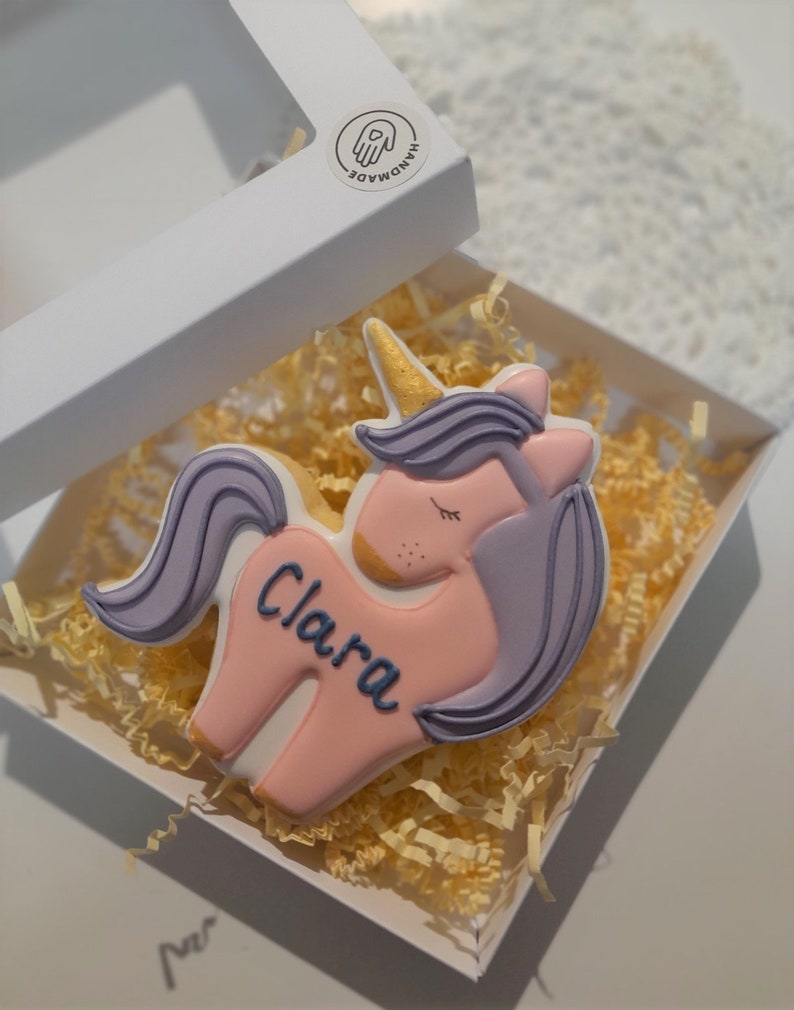 May include: A unicorn-shaped cookie with pink, purple, and gold details, personalized with the name "Clara" in blue. The cookie is presented in a white box with yellow paper filler, and a "HANDMADE" sticker.