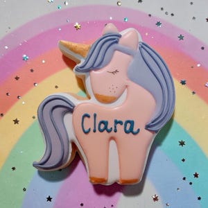 An unicorn-shaped biscuit with a pink body, purple mane and tail, and a golden horn. The biscuit has the name "Clara" written in blue.