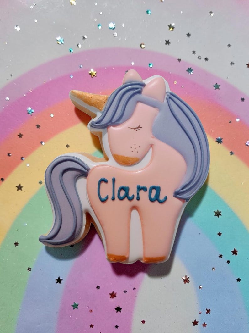 An unicorn-shaped biscuit with a pink body, purple mane and tail, and a golden horn. The biscuit has the name "Clara" written in blue.