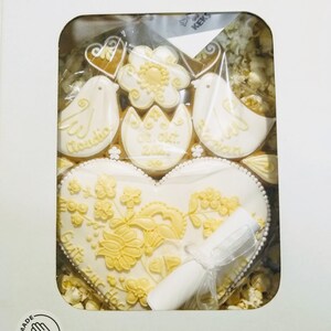 May include: A white heart-shaped cookie decorated with yellow floral designs and the text "Gute Reise zur Hochzeit". The cookie is surrounded by smaller cookies, including two doves, a flower, and a heart. The cookies are in a white box with a hand-drawn logo that says "Handmade".
