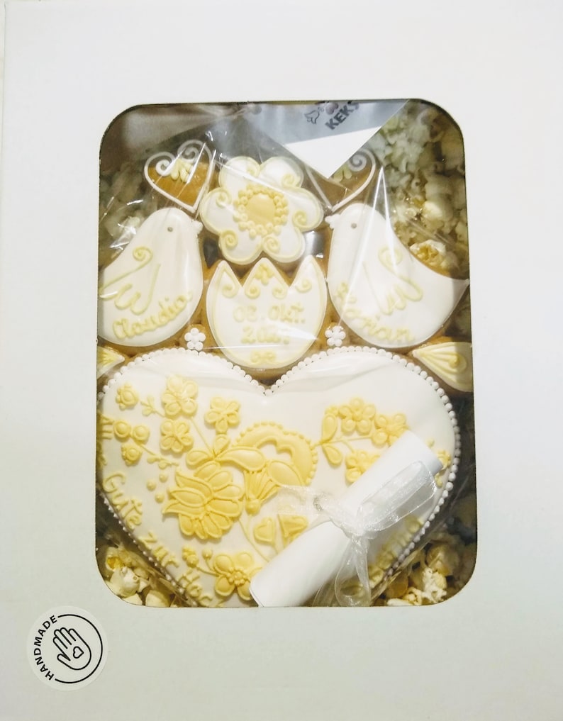 May include: A white heart-shaped cookie decorated with yellow floral designs and the text "Gute Reise zur Hochzeit". The cookie is surrounded by smaller cookies, including two doves, a flower, and a heart. The cookies are in a white box with a hand-drawn logo that says "Handmade".