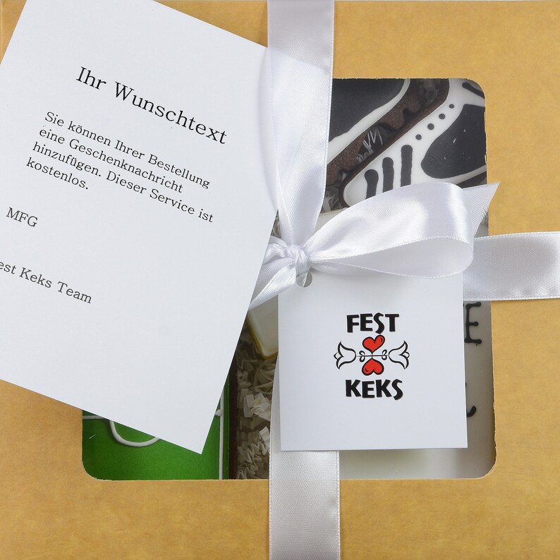 May include: A gift box tied with a white ribbon, featuring a tag with the text "FEST KEKS" and a heart design. The box contains a card with German text, including "Ihr Wunschtext". The box is brown with a window showing a decorated cookie.