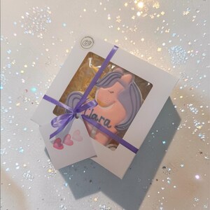 : A unicorn-shaped biscuit with a pink body, purple mane and tail, and a golden horn. The biscuit has the name "Clara" written in blue.