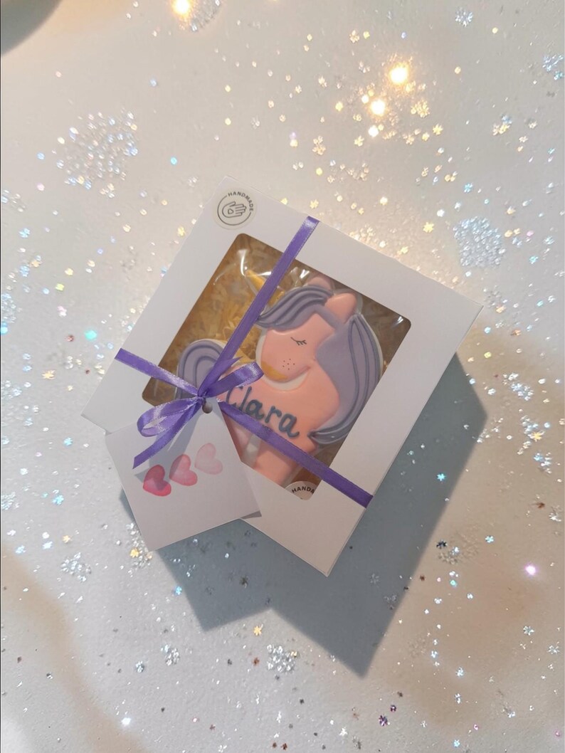 : A unicorn-shaped biscuit with a pink body, purple mane and tail, and a golden horn. The biscuit has the name "Clara" written in blue.