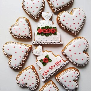 Puede incluir: Una vista a&eacute;rea de galletas de jengibre decoradas. Galletas en forma de coraz&oacute;n con glaseado blanco y lunares rojos rodean dos galletas en forma de pastel blancas. Un pastel tiene rosas rojas y el otro la fecha "5 10 19" en rojo.