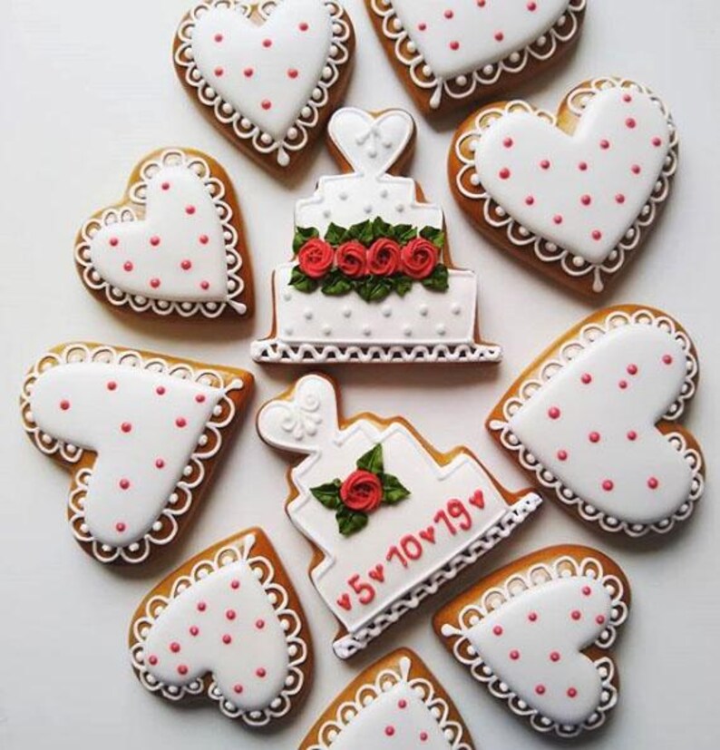 Puede incluir: Una vista a&eacute;rea de galletas de jengibre decoradas. Galletas en forma de coraz&oacute;n con glaseado blanco y lunares rojos rodean dos galletas en forma de pastel blancas. Un pastel tiene rosas rojas y el otro la fecha "5 10 19" en rojo.