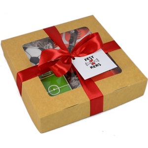 May include: A brown gift box tied with a red satin ribbon and bow. The box has a clear window showing soccer-themed cookies, including a soccer ball, a soccer field, and a pair of cleats. A tag with the text "Fest Keks" is attached.