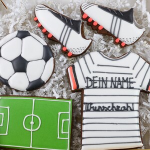 May include: A collection of soccer-themed cookies, including a soccer ball, a soccer field, two soccer cleats, and a jersey with the text "DEIN NAME" and "Wunschzahl". The cookies are decorated with white, black, green, and orange icing.