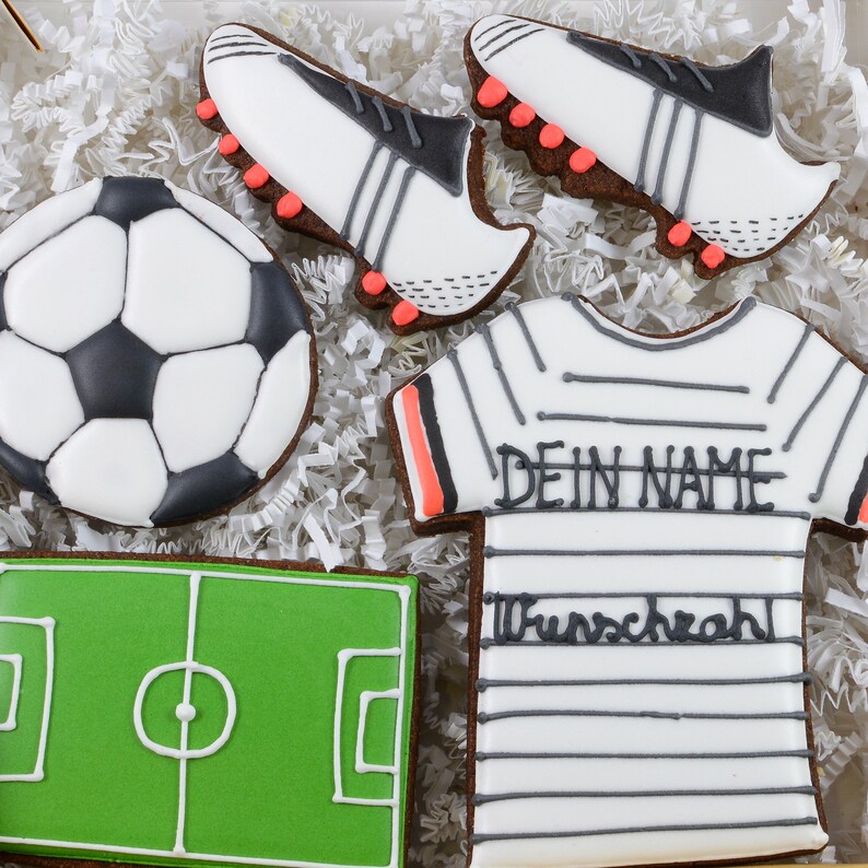 May include: A collection of soccer-themed cookies, including a soccer ball, a soccer field, two soccer cleats, and a jersey with the text "DEIN NAME" and "Wunschzahl". The cookies are decorated with white, black, green, and orange icing.