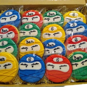 May include: A box of 12 decorated sugar cookies with a ninja theme. The cookies are decorated with green, red, blue, and yellow icing. The cookies have the number 6 and the name Ben written on them.