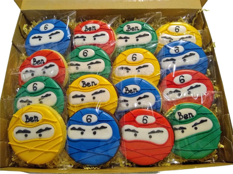 May include: A box of 12 decorated sugar cookies with a ninja theme. The cookies are decorated with green, red, blue, and yellow icing. The cookies have the number 6 and the name Ben written on them.
