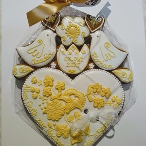 May include: A white gingerbread heart with yellow floral designs and the text "Alles Gute zur Hochzeit" and "Marlis & Bernd". Two white doves with the names "Claudia" and "Florian" are on top of the heart. The date "08. Okt. 2021" is written on a yellow flower.