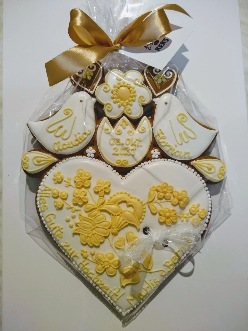 May include: A white gingerbread heart with yellow floral designs and the text "Alles Gute zur Hochzeit" and "Marlis & Bernd". Two white doves with the names "Claudia" and "Florian" are on top of the heart. The date "08. Okt. 2021" is written on a yellow flower.