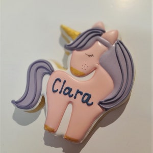 May include: A unicorn-shaped cookie with a pink body, lavender mane and tail, and a golden horn. The name "Clara" is written in blue script across the body. The cookie is decorated with white icing and gold accents.