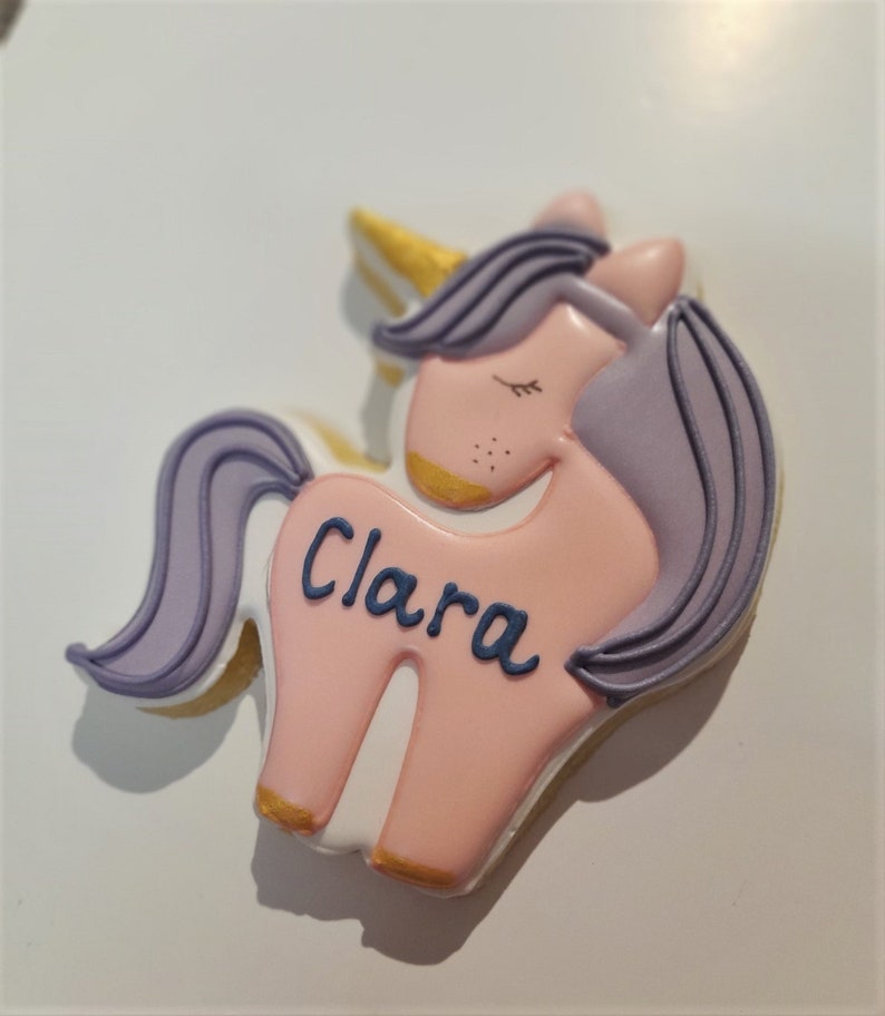 May include: A unicorn-shaped cookie with a pink body, lavender mane and tail, and a golden horn. The name "Clara" is written in blue script across the body. The cookie is decorated with white icing and gold accents.