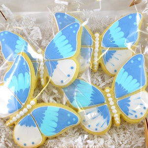 Op de afbeelding: Vier blauwe en witte vlinderkoekjes versierd met witte parel-achtige accenten. De koekjes zijn verpakt in een doorzichtige plastic zak en omgeven door wit gescheurd papier.